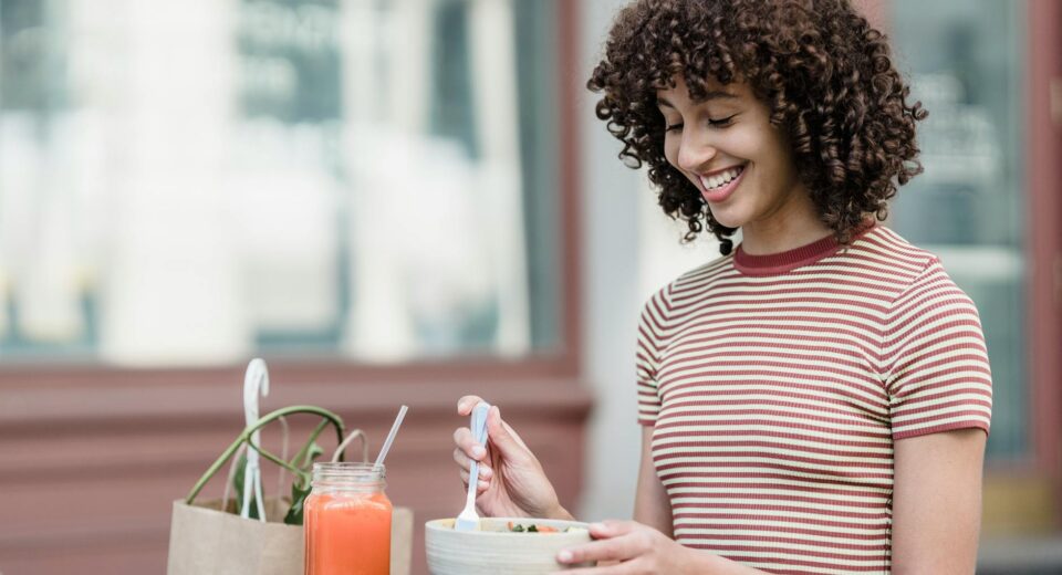 Cheerful young female with bowl of yummy food and jar of refreshing beverage sitting at table in urban cafeteria