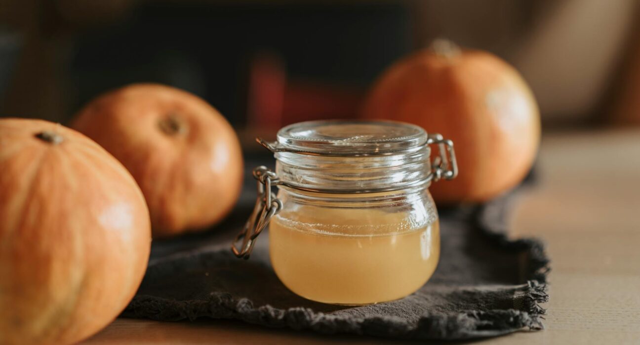 A cozy still life with homemade apple cider in a glass jar surrounded by pumpkins on a wooden table.
