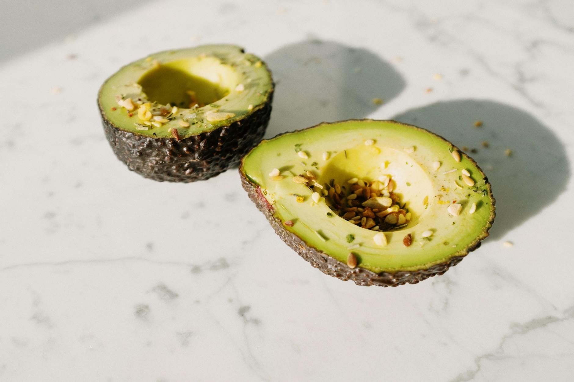 From above of green halved avocado without kernel sprinkled with various seeds placed on white marble desk served for healthy vegan breakfast preparation