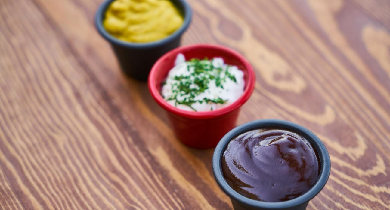 A close-up of three sauces in cups on a wooden surface, arranged in line.