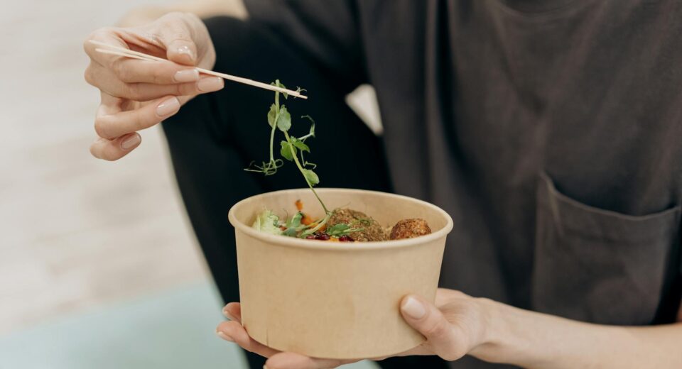 A woman holding a paper bowl filled with a healthy vegetarian meal in an indoor setting, promoting wellness.