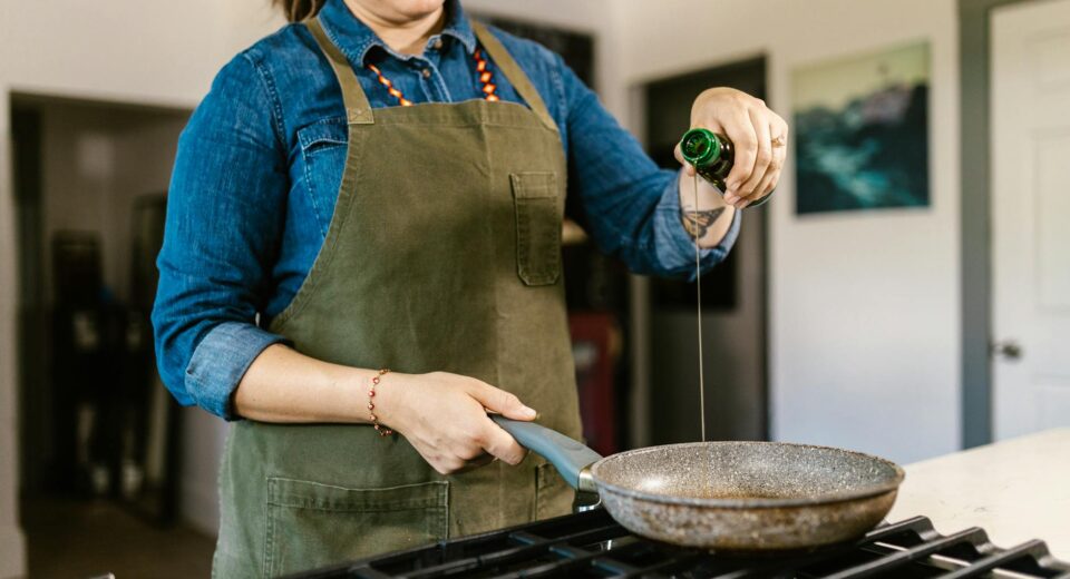 A home chef in an apron pours olive oil into a frying pan on a stove.