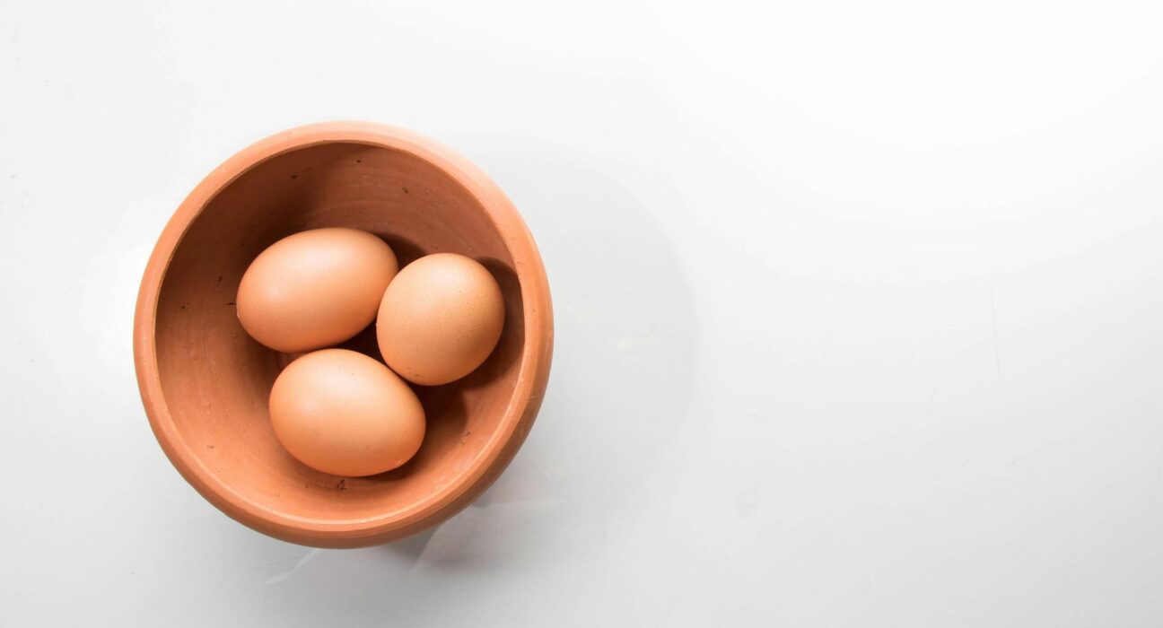 A top-down view of three brown eggs in a brown ceramic bowl on a white background.