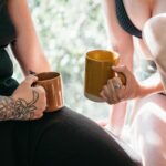 Two women enjoying coffee indoors, showcasing tattoos and rings in natural light.