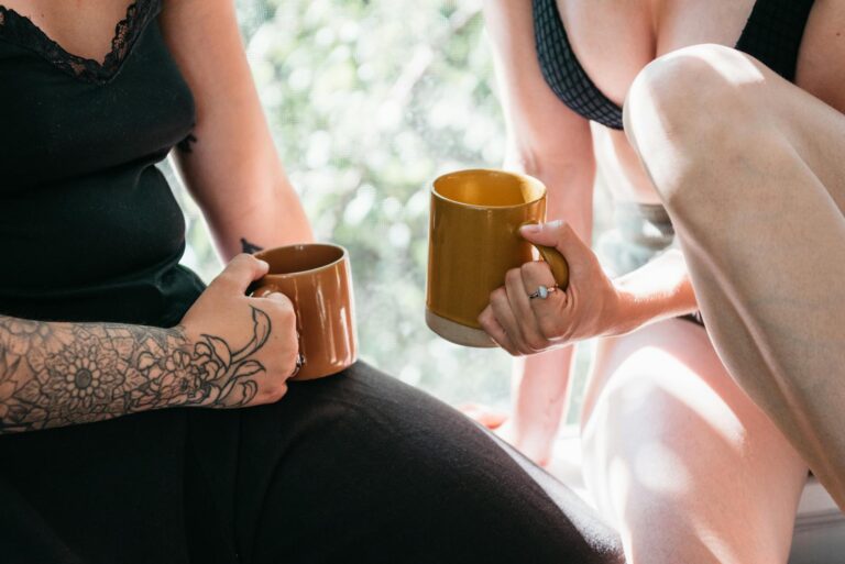 Two women enjoying coffee indoors, showcasing tattoos and rings in natural light.