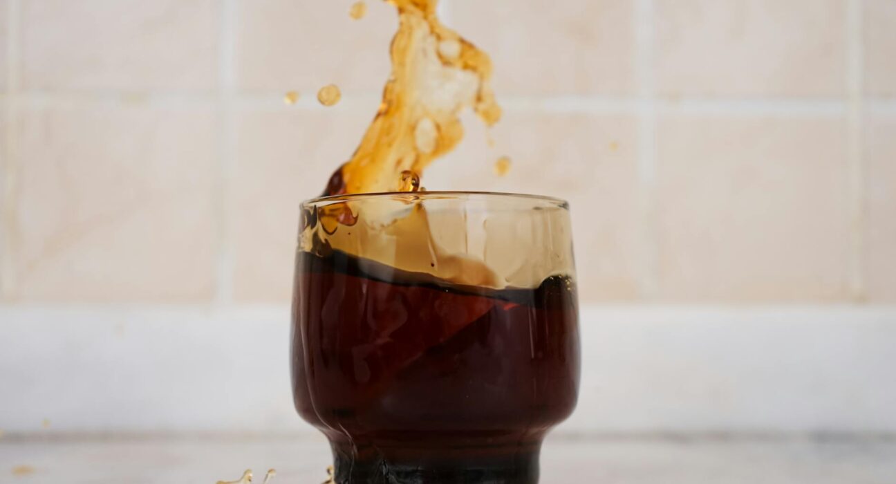 Energetic splash of coffee in a clear glass against a tiled background, capturing motion and excitement.