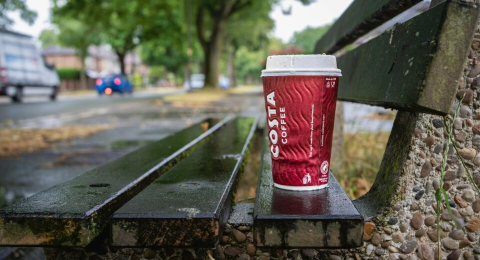 Close-up of a coffee cup on a wet bench, vibrant street scene after rain in Urmston.