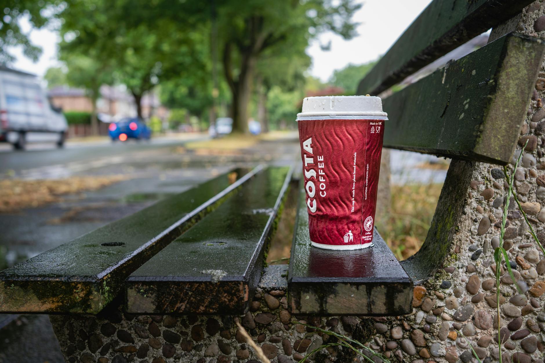 Close-up of a coffee cup on a wet bench, vibrant street scene after rain in Urmston.