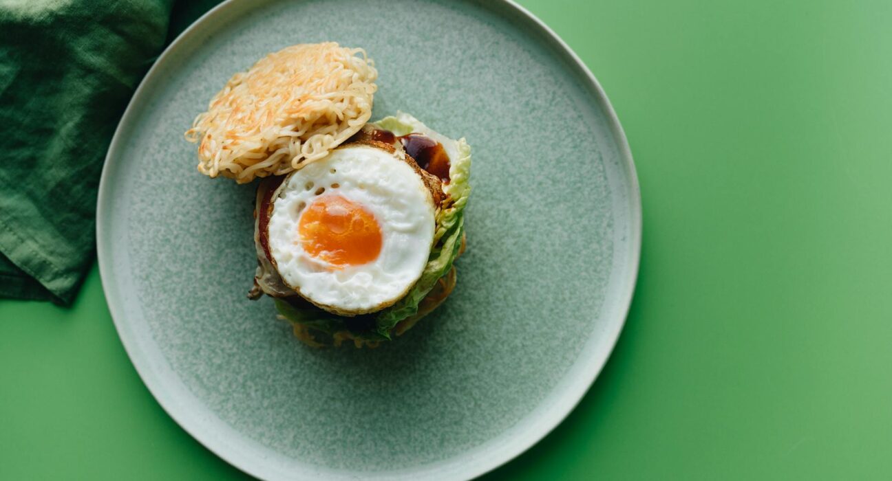 Delicious ramen burger topped with a fried egg on a green ceramic plate. Perfect for food photography.
