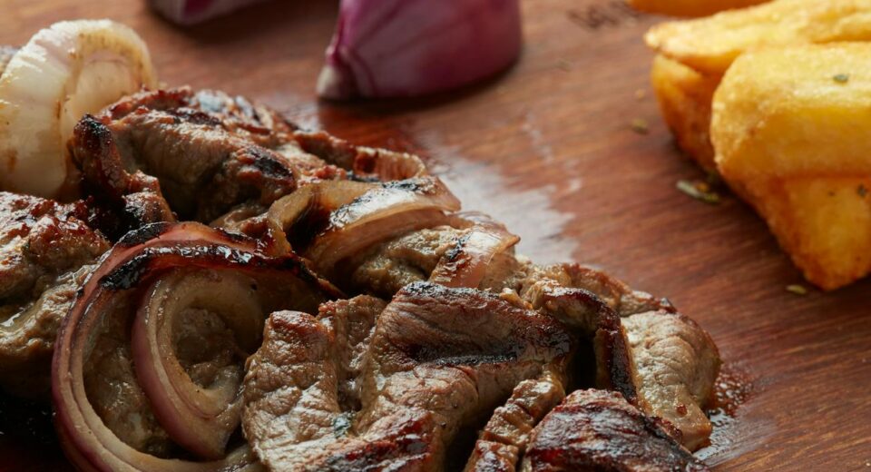 Delicious grilled steak with red onion and fried yuca on a wooden board, offering a savory meal.