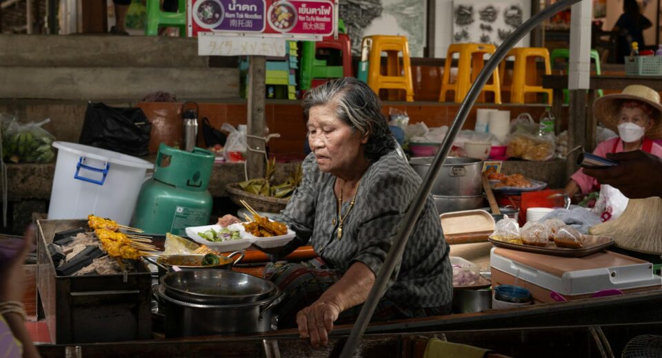Elderly woman cooking traditional Thai dishes at a bustling Bangkok street market.