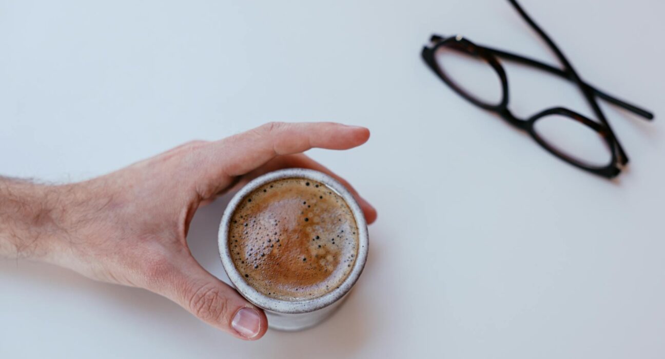 A close-up of a hand holding a cappuccino mug on a white table with glasses nearby.