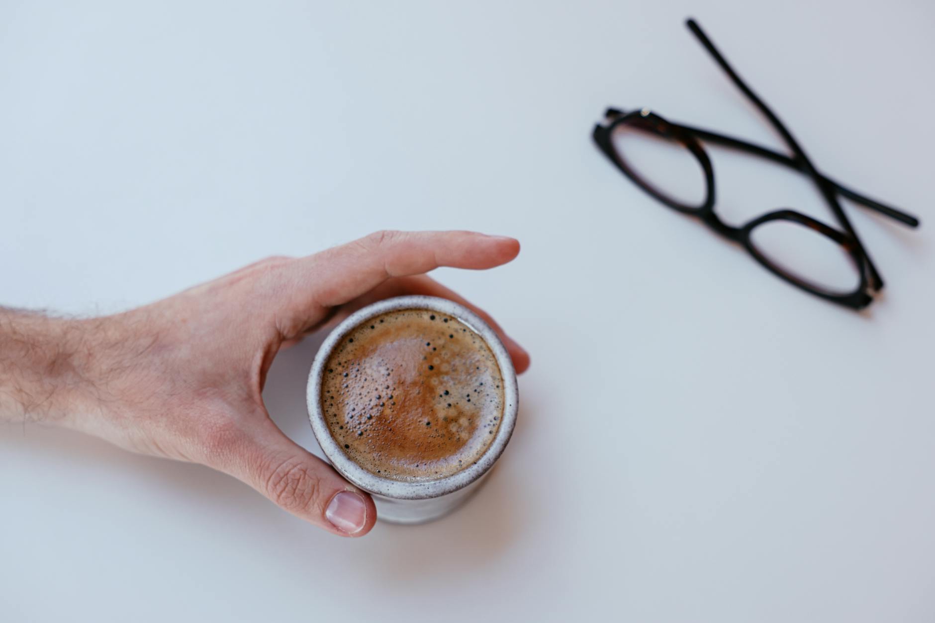 A close-up of a hand holding a cappuccino mug on a white table with glasses nearby.