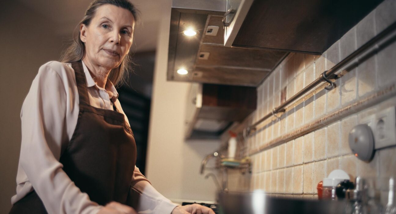 A mature woman in an apron stands confidently in a modern kitchen interior.