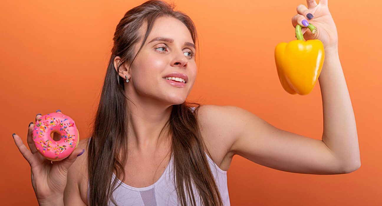 Woman in sports bra juggling choice between a donut and bell pepper on orange background.