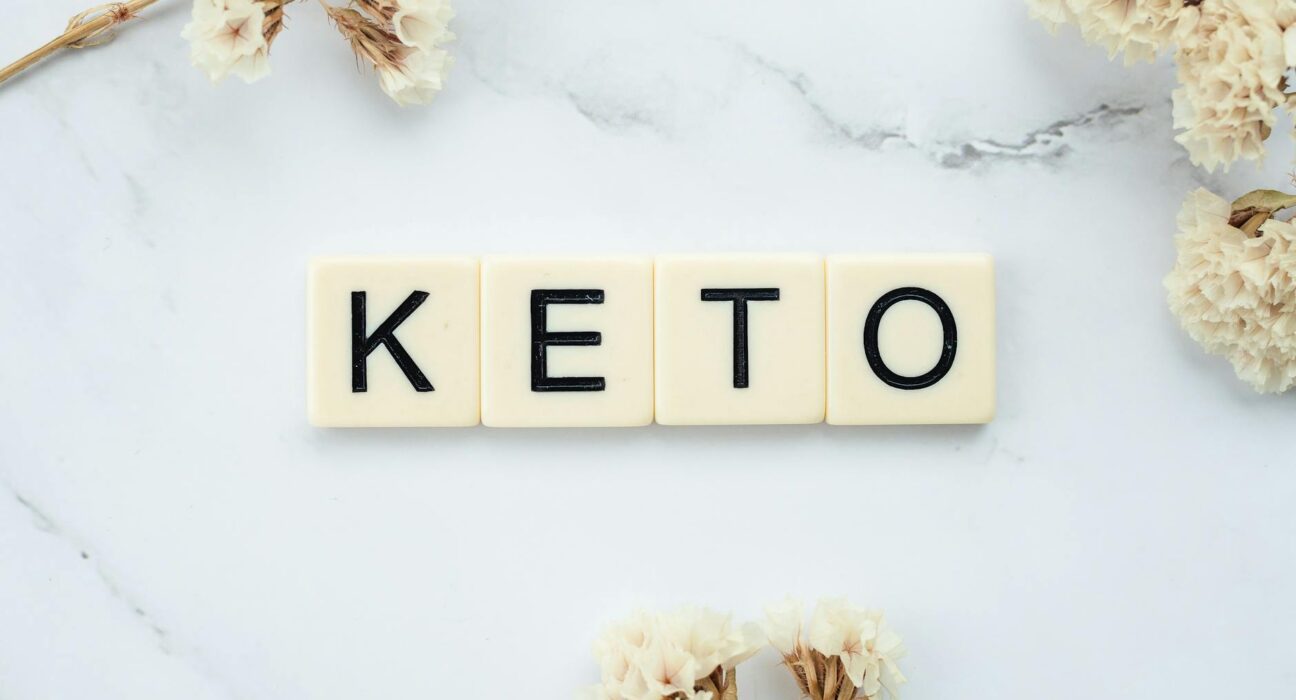 Scrabble tiles spelling 'Keto' on a marble surface with white flowers, symbolizing a ketogenic lifestyle.