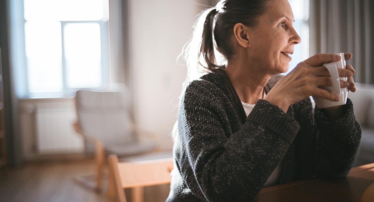 A senior woman in a cozy sweater enjoys a warm drink while sitting indoors, captured in soft light.