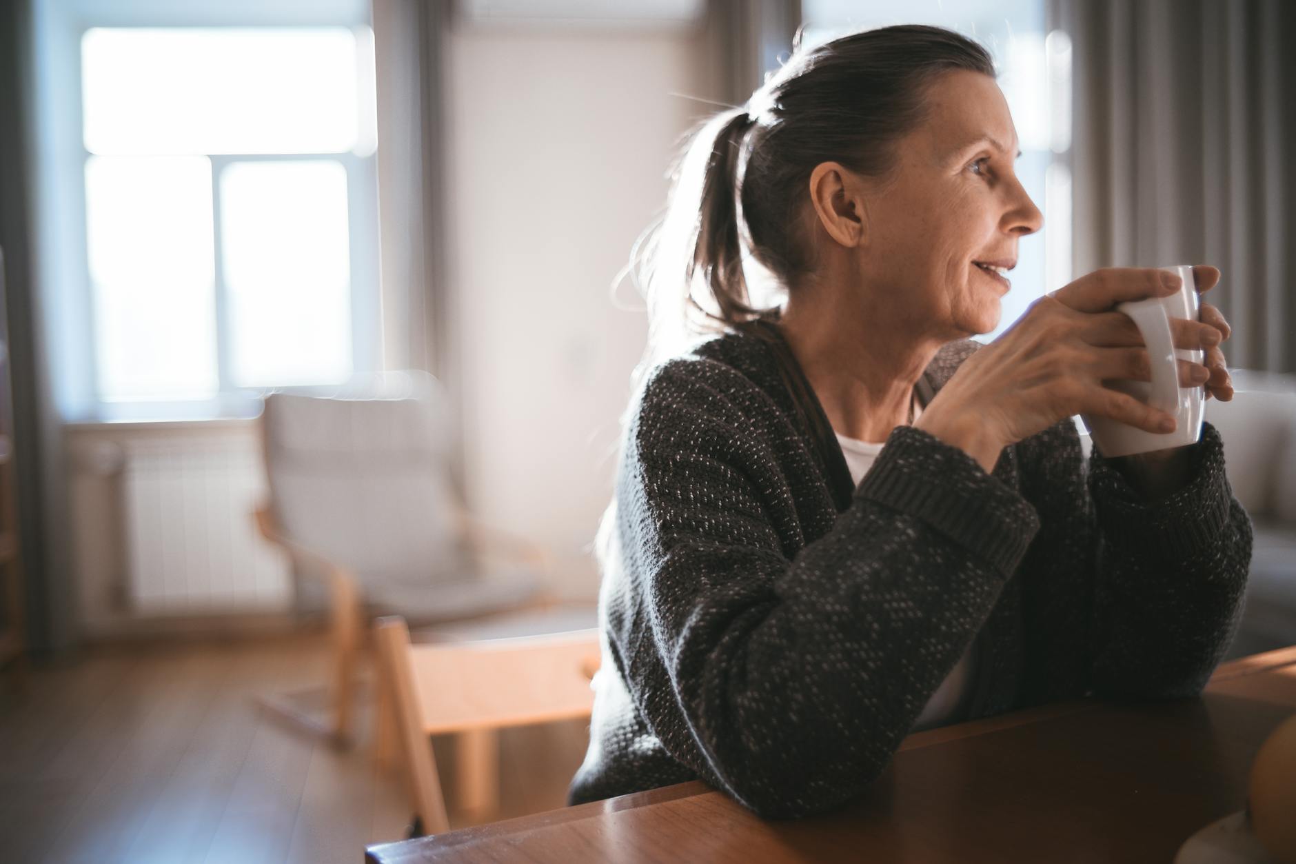 A senior woman in a cozy sweater enjoys a warm drink while sitting indoors, captured in soft light.