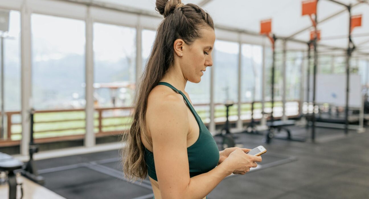 Female athlete in gym looking at phone, dressed in activewear for training.