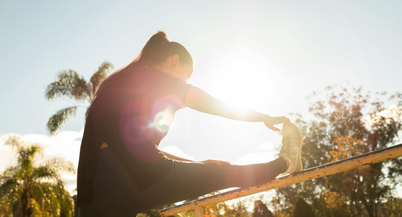 A woman stretches on a bar in a sunlit park, surrounded by trees and a clear sky.