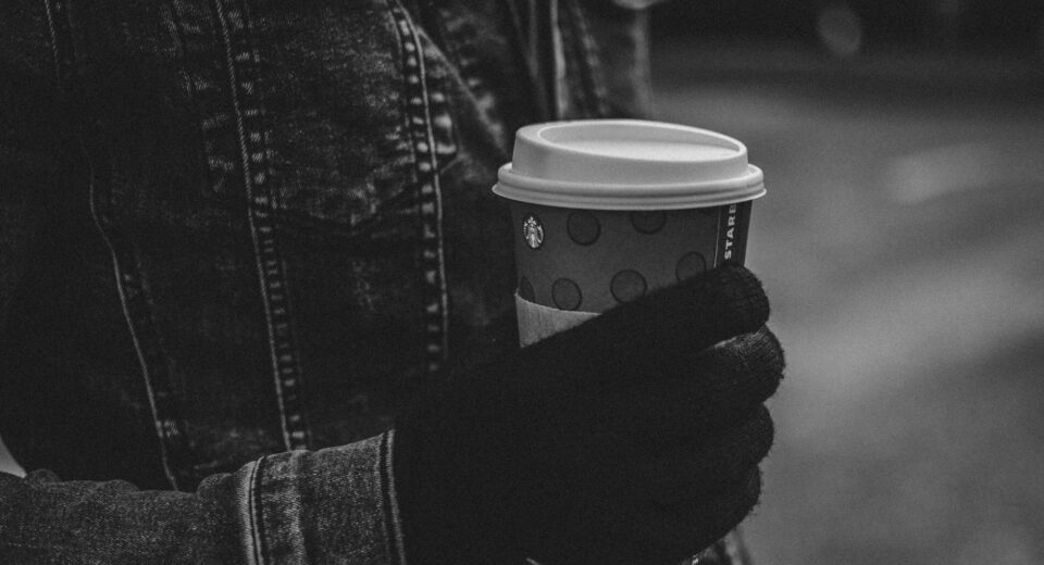 A person in a denim jacket and gloves holding a coffee cup outdoors.