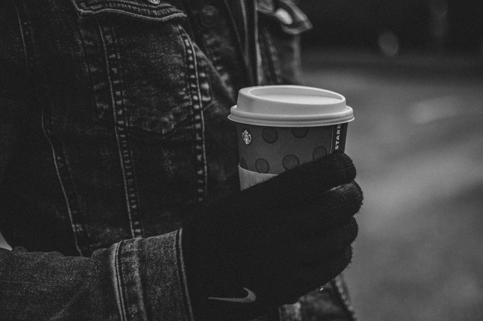 A person in a denim jacket and gloves holding a coffee cup outdoors.