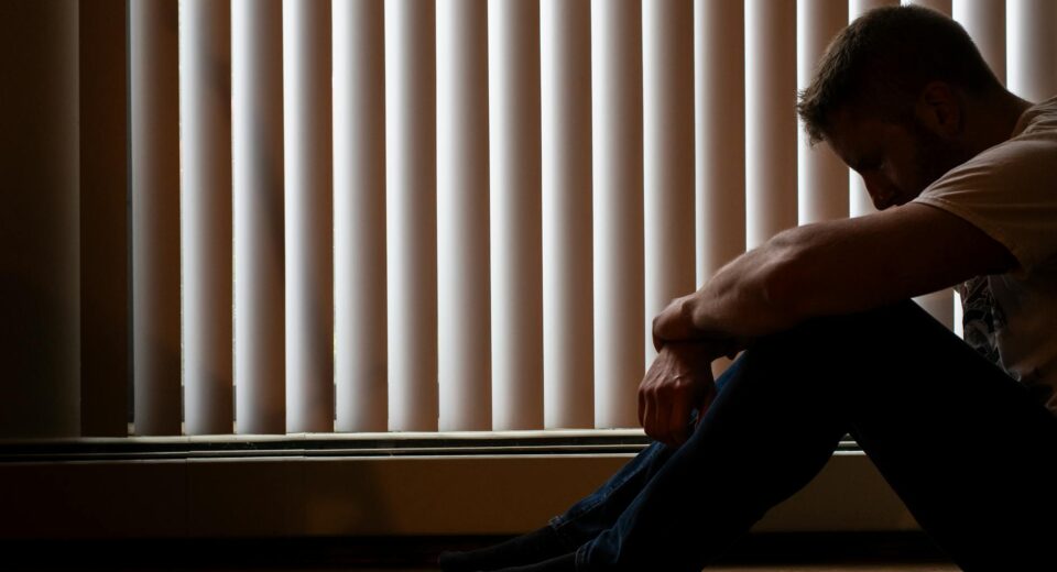 A man sits silhouetted against window blinds, conveying solitude and introspection.