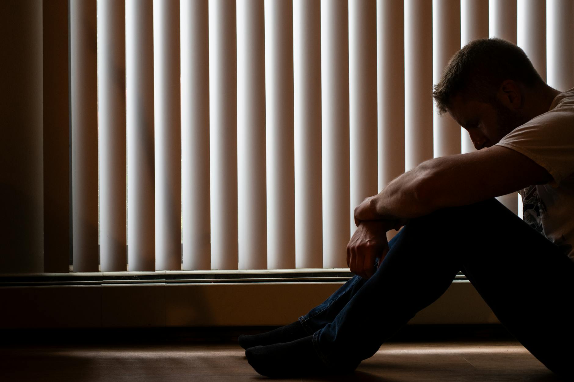 A man sits silhouetted against window blinds, conveying solitude and introspection.