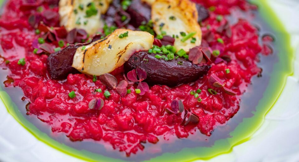 A colorful beetroot risotto topped with grilled halloumi and fresh herbs on a white plate.