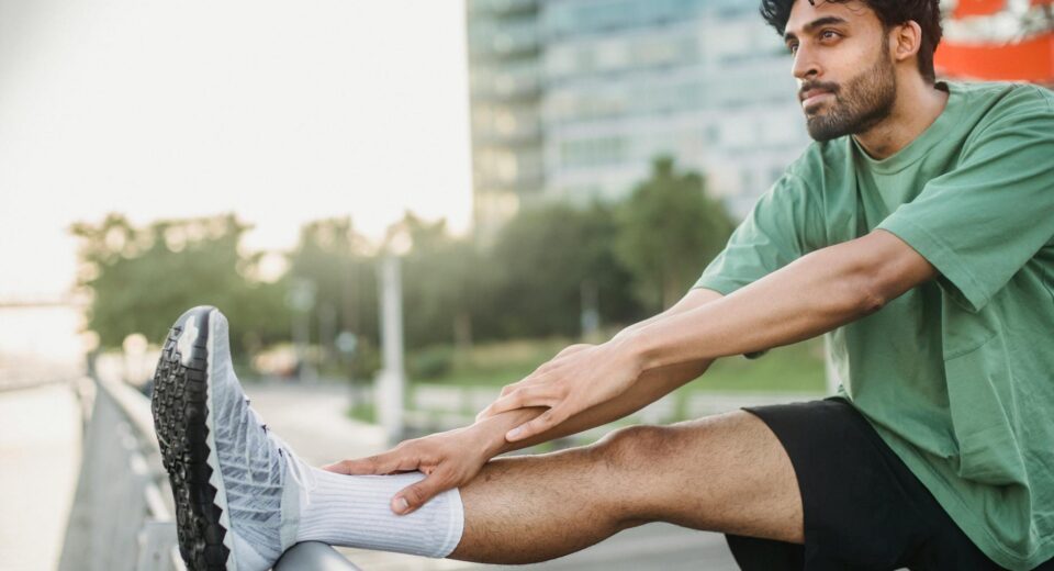 A man in a green shirt stretches his leg outdoors near a modern building, promoting a healthy lifestyle.