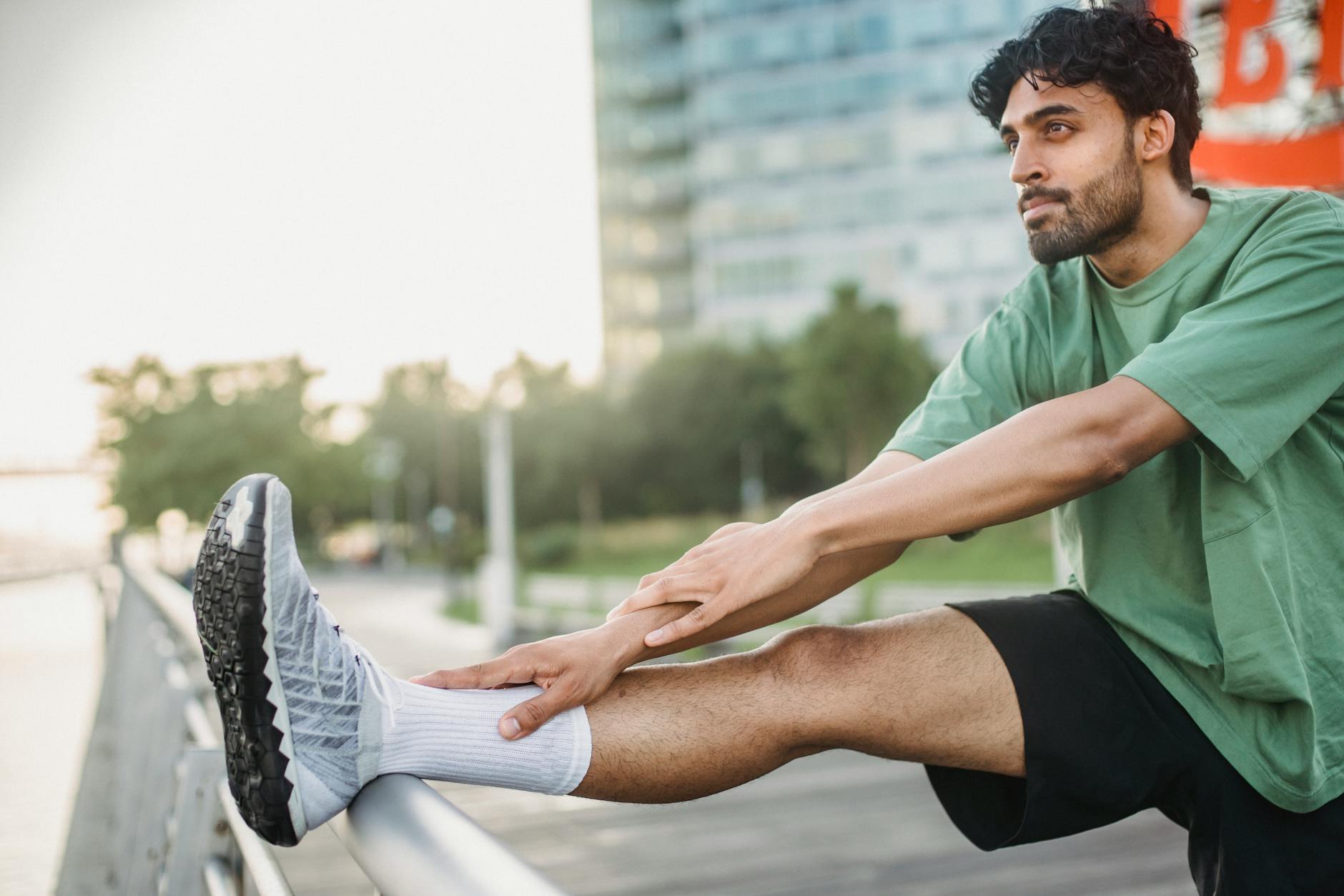 A man in a green shirt stretches his leg outdoors near a modern building, promoting a healthy lifestyle.