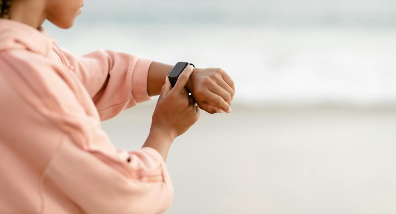 Woman wearing pink hoodie checks smartwatch by the ocean, focused on time tracking.