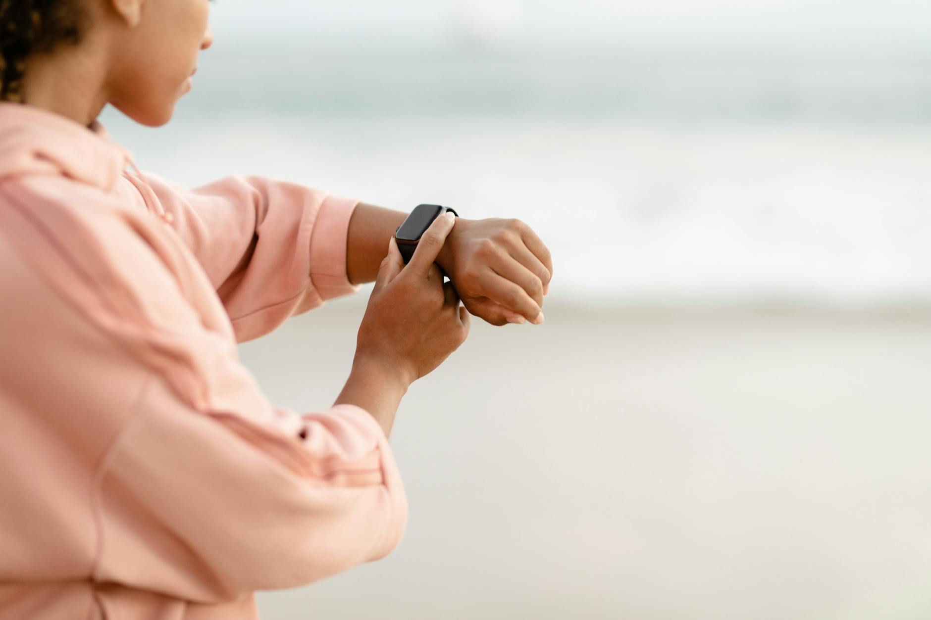 Woman wearing pink hoodie checks smartwatch by the ocean, focused on time tracking.