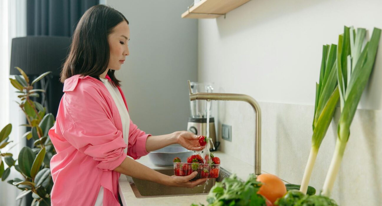 Asian woman washes strawberries at kitchen sink, promoting healthy living.