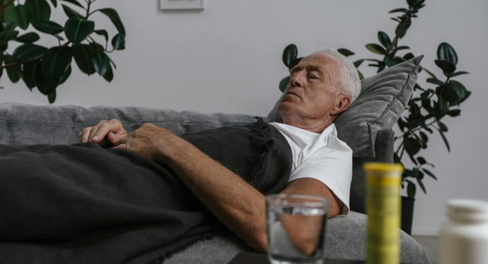 An elderly man sleeping on a couch with a blanket and glass of water nearby.