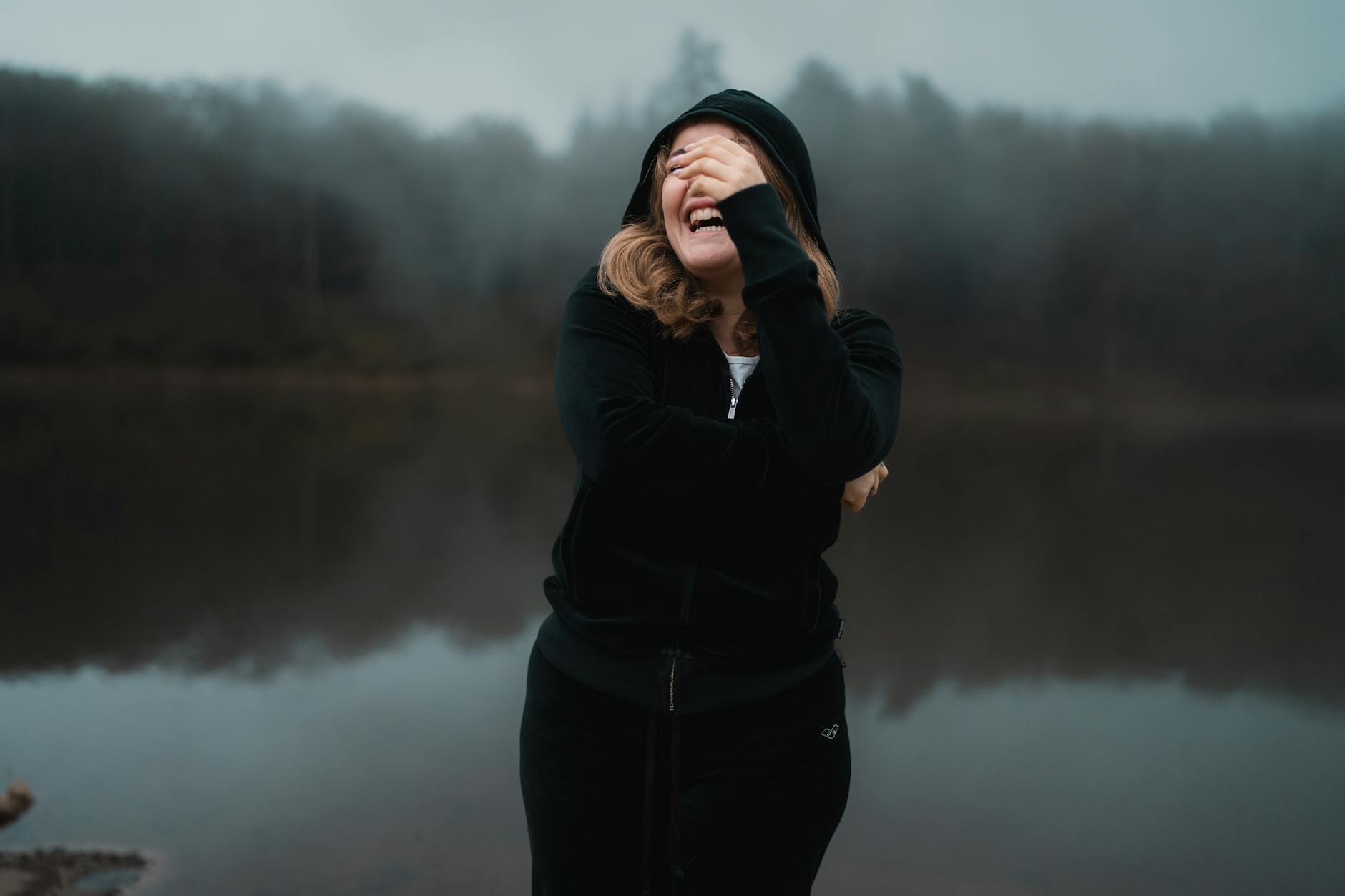 A woman laughing near a serene, foggy lake in autumn, capturing natural joy.