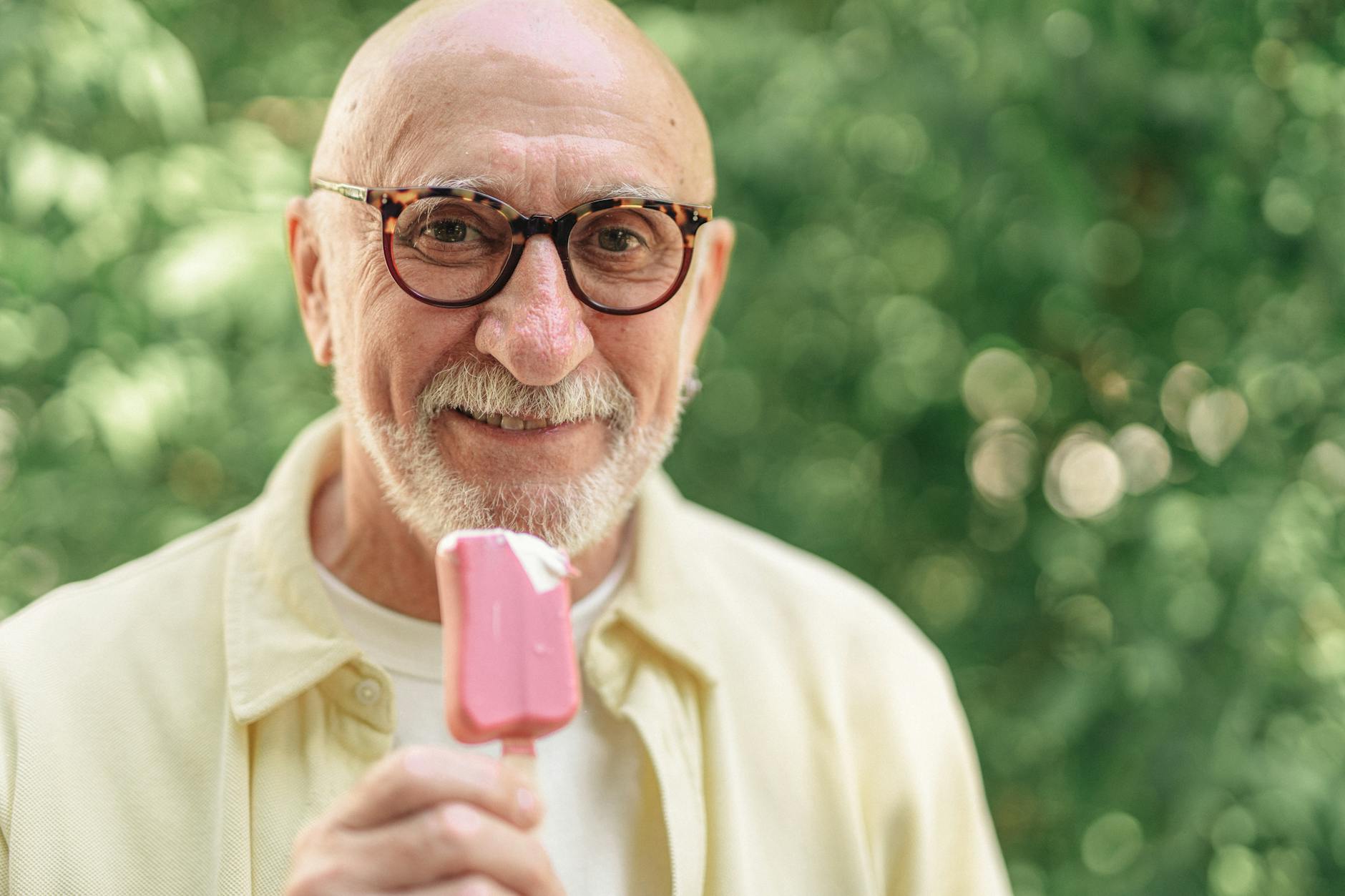 Elderly man wearing glasses and holding a pink popsicle, enjoying a sunny day outside.