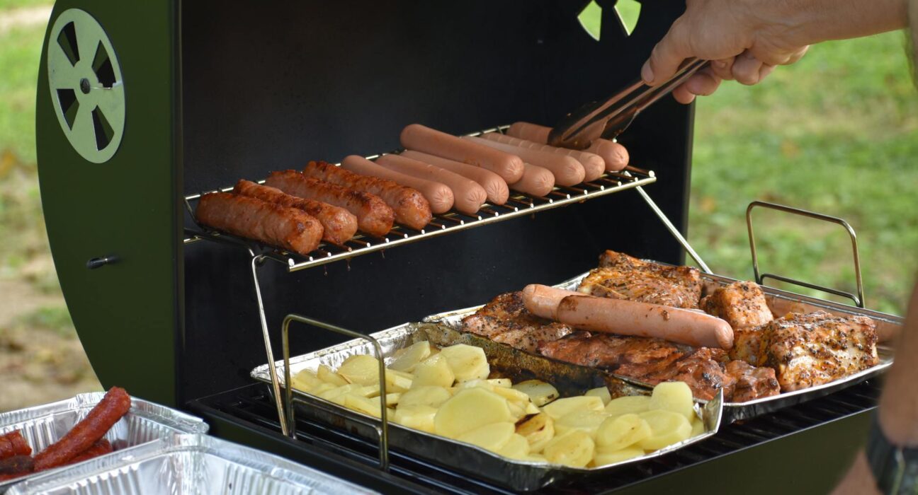 Outdoor barbecue with hotdogs, sausages, potatoes, and chicken grilling on a sunny day.