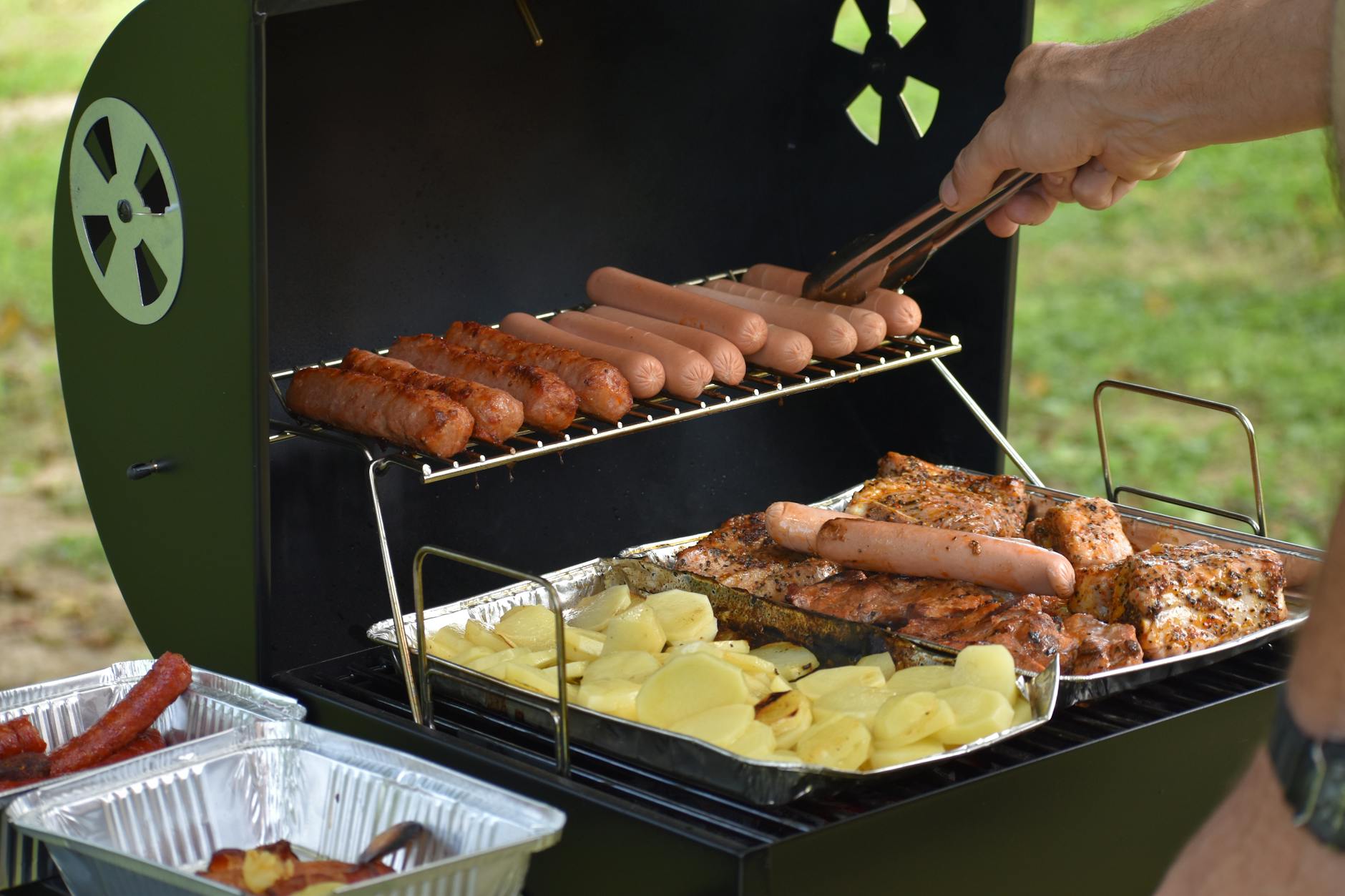 Outdoor barbecue with hotdogs, sausages, potatoes, and chicken grilling on a sunny day.