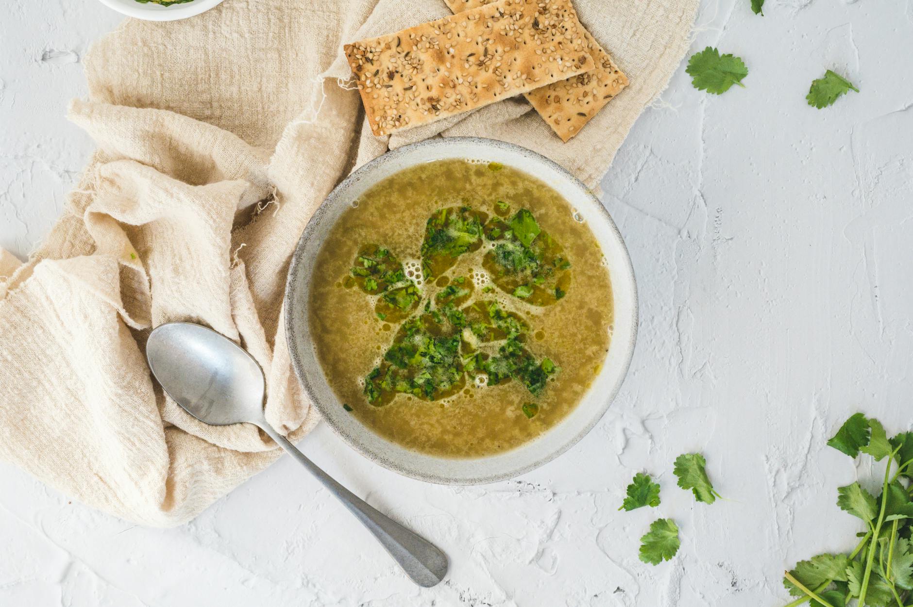 A bowl of fresh herb soup served with crackers and a spoon on a light background.