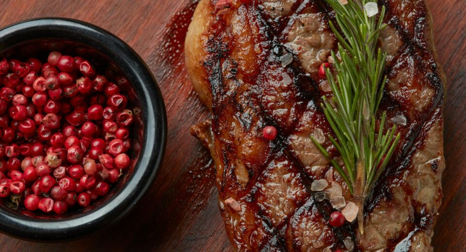 Close-up of grilled steak garnished with rosemary and pink peppercorns, enhancing its flavor.