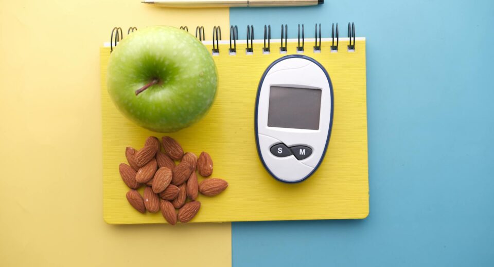 Flat lay of diabetes tools, green apple, almonds, and notebook for health planning.