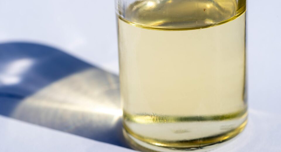 A close-up image of a glass bottle with liquid, showcasing light reflection and shadow on a white surface.