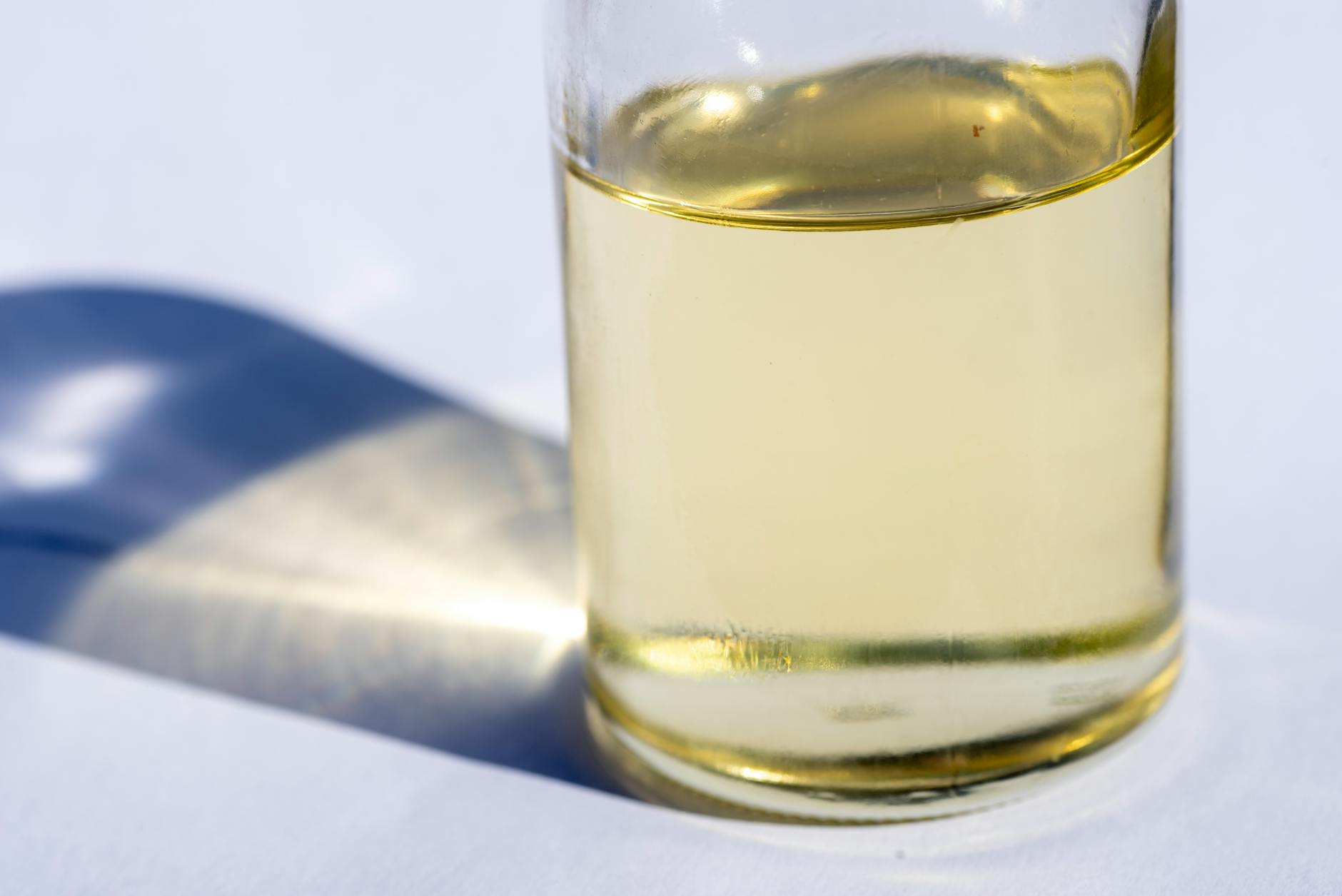 A close-up image of a glass bottle with liquid, showcasing light reflection and shadow on a white surface.