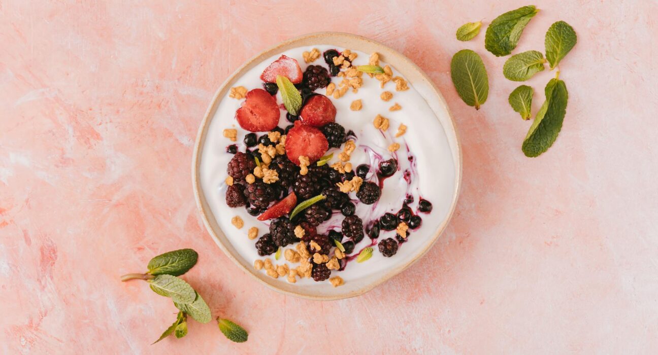 Top view of a vibrant yogurt bowl with mixed berries, granola, and mint leaves on a pink background.