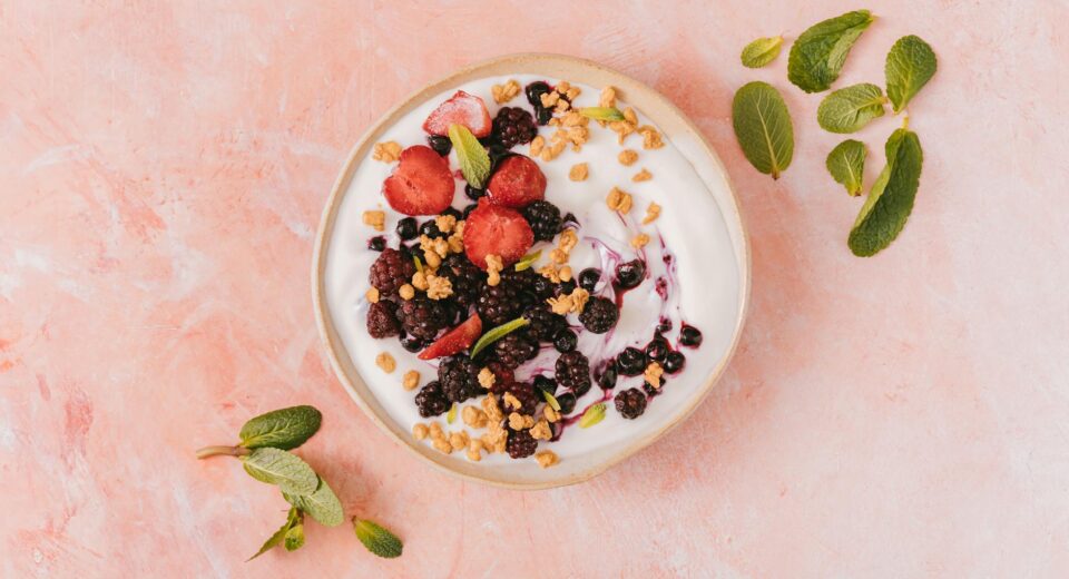 Top view of a vibrant yogurt bowl with mixed berries, granola, and mint leaves on a pink background.