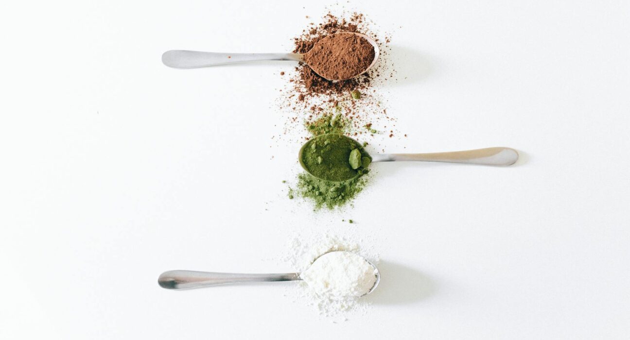 Top view of colorful assorted powders on spoons against white background.