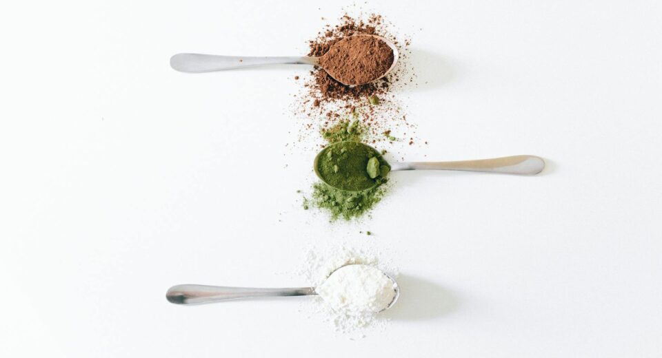 Top view of colorful assorted powders on spoons against white background.