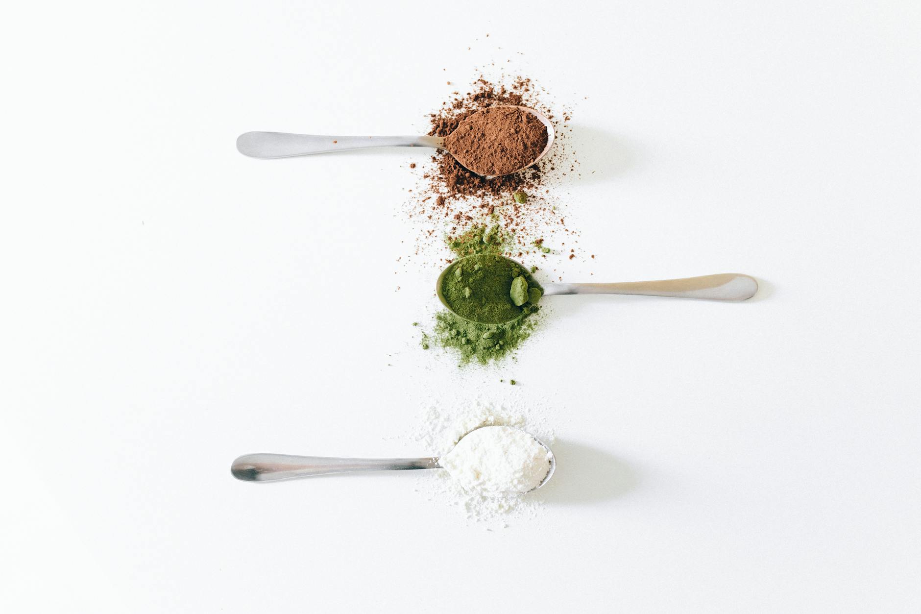Top view of colorful assorted powders on spoons against white background.