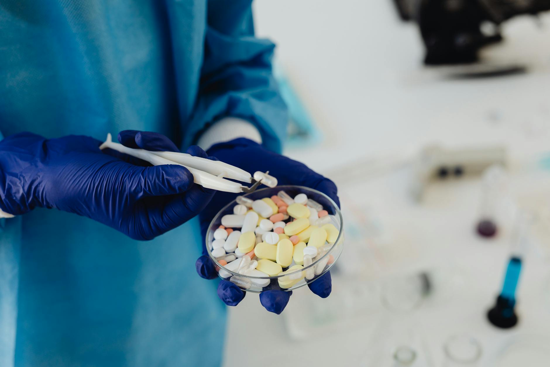Scientist in gloves using tweezers to handle assorted pills in a petri dish.